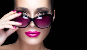 Beautiful fashion model girl with hand on stylish oversized pink sunglasses looking at camera. Bright makeup and manicure. High fashion portrait isolated on black background. Beauty and fashion concept.